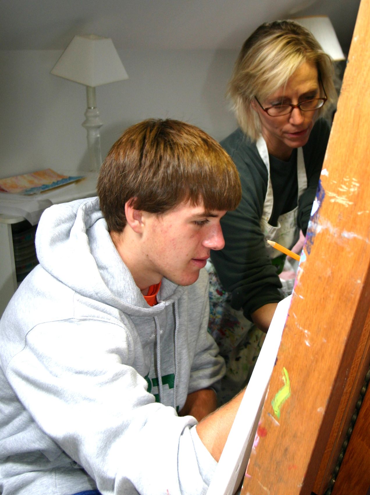 An instructor guiding a teenage student painting at the easel, one-on-one